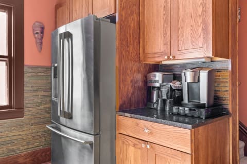 A kitchen corner featuring a stainless steel refrigerator, coffee maker, and wooden cabinets against a vibrant peach wall.