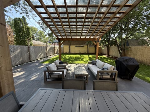A backyard with a wooden pergola, outdoor seating, a coffee table, and a grill on a sunny day.