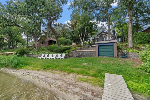 Lakeside view featuring white Adirondack chairs on the grass, a wooden house, and tall trees.