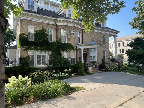 Elegant brick mansion with climbing ivy and bright garden flowers, surrounded by mature trees.
