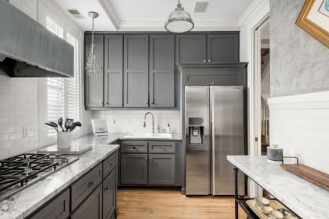 A stylish kitchen with gray cabinets, marble countertops, stainless steel appliances, and natural light filtering through the windows.