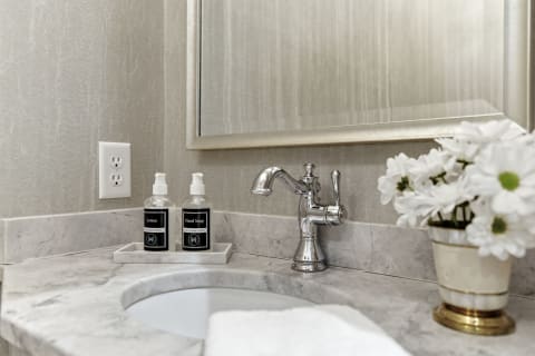 A stylish bathroom with marble countertops, a chrome faucet, hand soap and lotion bottles, and white flowers in a vase.