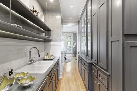 A stylish gourmet galley kitchen with gray cabinetry, white tile, and warm hardwood flooring, showcasing open shelves and elegant décor.
