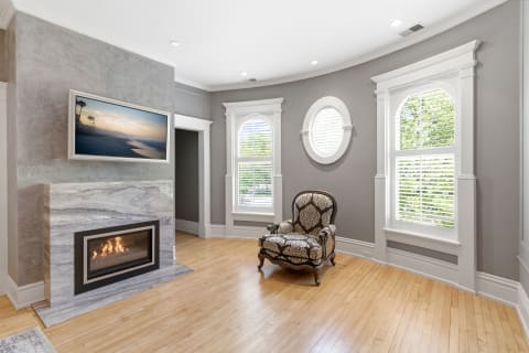 A cozy room with a marble fireplace, ornate armchair, and beautiful light from large windows.