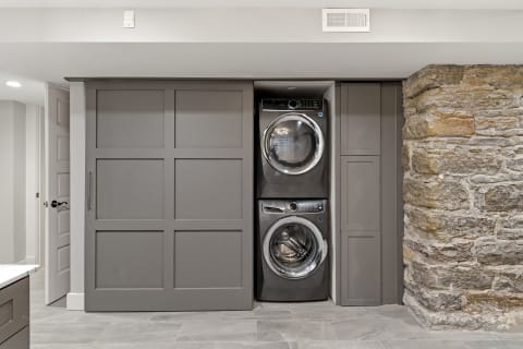 Stacked washer and dryer in custom cabinetry beside a stone wall, featuring gray panels and tile flooring.