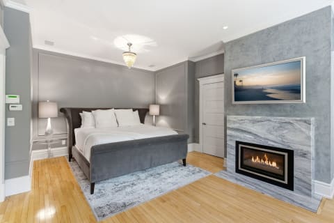 A serene bedroom with a plush king-sized bed and a modern fireplace, styled in soft grey tones.