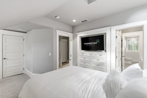 Modern bedroom with white linens, wall-mounted TV, and an open door to an ensuite bathroom.