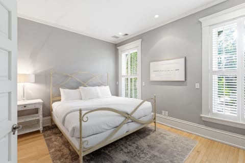 A calming bedroom featuring a wrought-iron bed with white linens, soft gray walls, and a bedside lamp beside a vintage table.