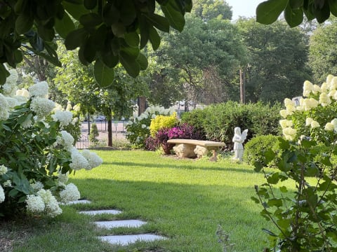 A beautifully landscaped garden with white hydrangeas, a stone bench, and an angel statue amidst lush greenery.