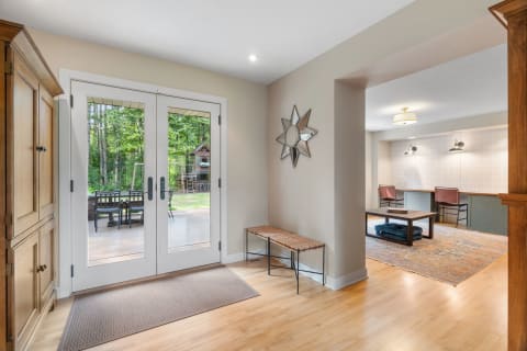 An entryway with glass doors, a wooden cupboard, a woven bench, and an office space in the background.